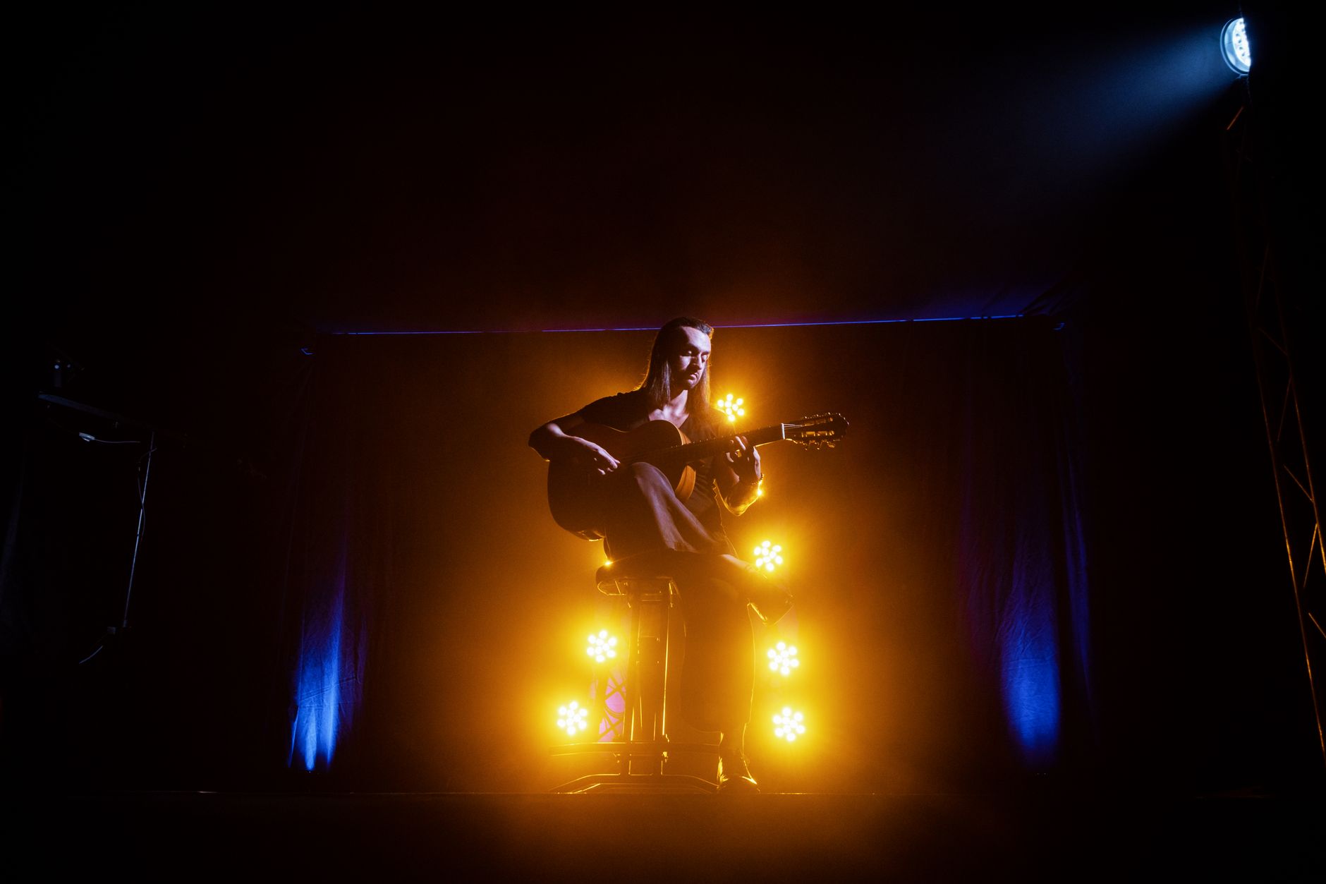 A musician playing an acoustic guitar on stage, illuminated by dramatic yellow and blue lights.