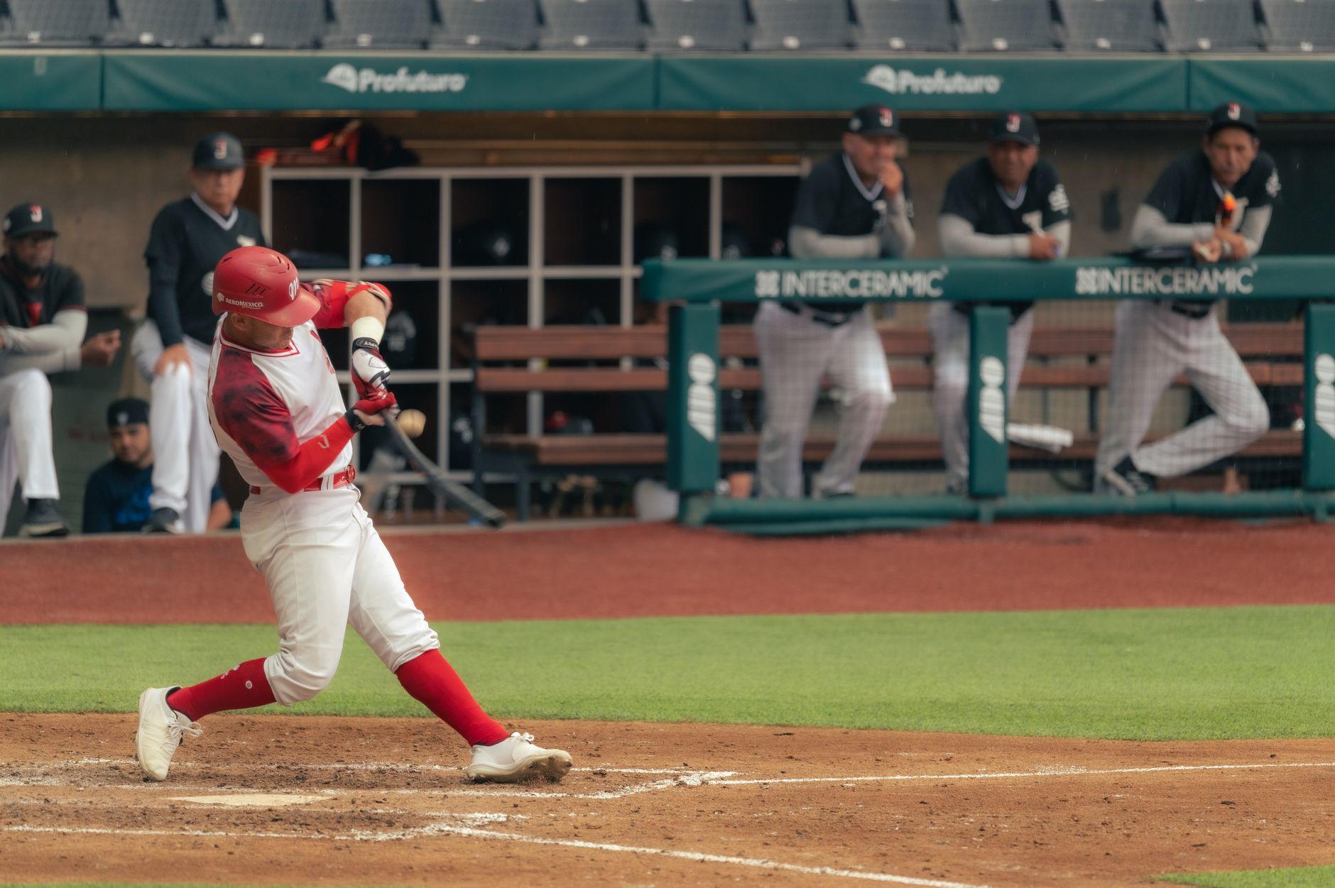 Baseball player swinging at a pitch during a game with coaches watching from the dugout.